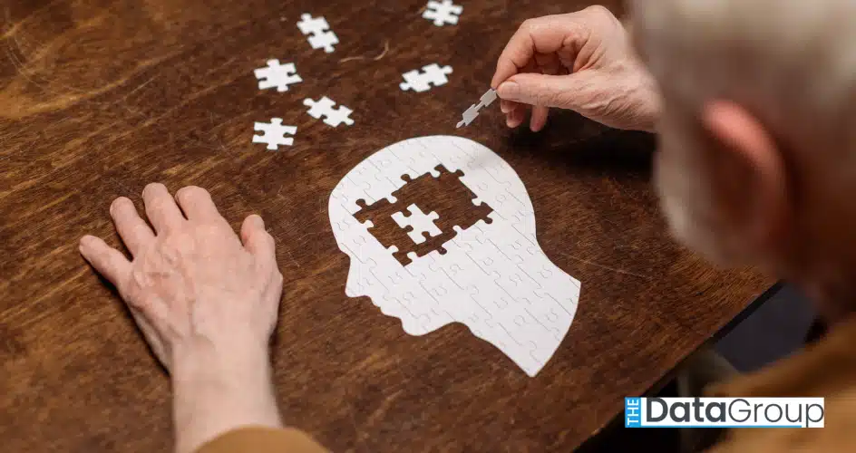 A mature adult is seen assembling a white puzzle in the shape of a human head. Set on a wooden table, the brain-teaser consists of multiple pieces, some of which are still unassembled. The photo conveys a sense of challenge and determination. This image would be perfect for projects centered on mental health, wellness, and cognitive function. It would also work well for campaigns related to dementia or memory loss.